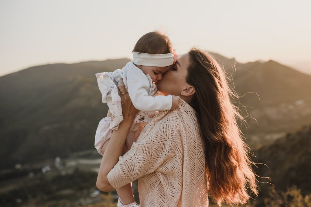 A mother gently kissing her baby while standing outdoors with a mountainous backdrop.